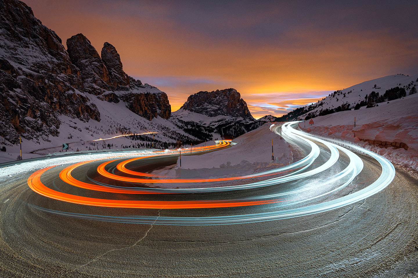 Car trail lights on bends of Gardena Pass road at sunset with Sassolungo on background in winter, Dolomites, South Tyrol, Italy